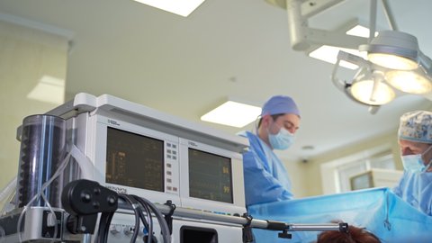 KYIV, UKRAINE - August 2021: Breathing ventilator of the anesthesia machine in motion. Black screen showing the vital signs of a patient. Doctors perform surgery at backdrop. Low angle view.