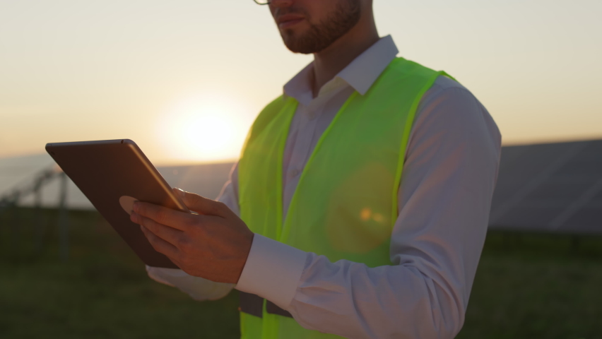 Close up of male engineer in safety vest using digital tablet while working on station with solar panels. Caucasian man standing outdoors with sunset on background. Royalty-Free Stock Footage #1088426827