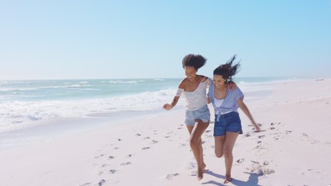 Smiling young black woman having fun at beach with her best friend. Latin hispanic young women running on seashore barefoot during vacation. Cheerful friends enjoying at sea on a bright sunny day.