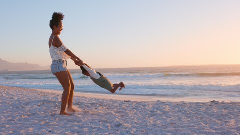 Playful young mother with laughing daughter fooling around at tropical beach during sunset. Young mother having fun while swinging her little black girl at sunset. Mother and daughter playing together