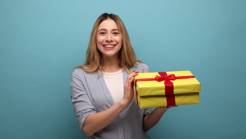 Portrait of happy woman opening gift box, looking inside with happy expression, being glad to get such present, wearing striped shirt. Indoor studio shot isolated on blue background. | Shutterstock HD Video #1088428381