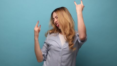 Portrait of overjoyed woman showing rock and roll hand sign, screaming and gesturing to heavy metal, rock music, wearing striped shirt. Indoor studio shot isolated on blue background.