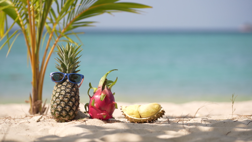 Ripe attractive pineapple and pink dragon fruit in sunglasses and fresh durian on the sand tropical beach against turquoise sea water, Thailand. Summer vacation concept
