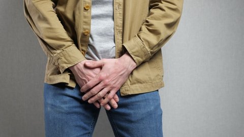 Man in jacket suffers from urinary incontinence trying to delay going to toilet. Male person has health problems standing on grey background closeup
