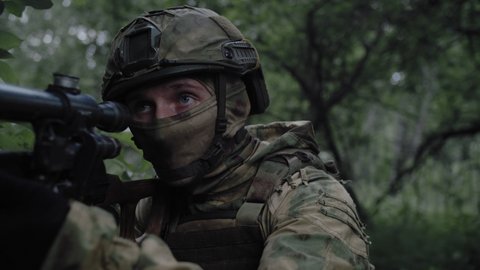 Close-up, an armed soldier with a sniper rifle, in a dense forest, protects the front line, in combat readiness aims at the enemy through an optical sight. Equipped soldier in action