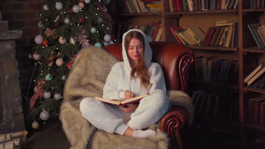 Christmas. Woman reading book sitting cozy leather armchair with Christmas tree in background . Beautiful young caucasian female resting home reads book dressed warm pajamas jumpsuit