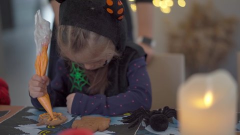 Little girl decorating icing handmade Halloween cookies sitting on the table dressed Halloween carnival costume with horns . Halloween party in big family. High quality 4k footage