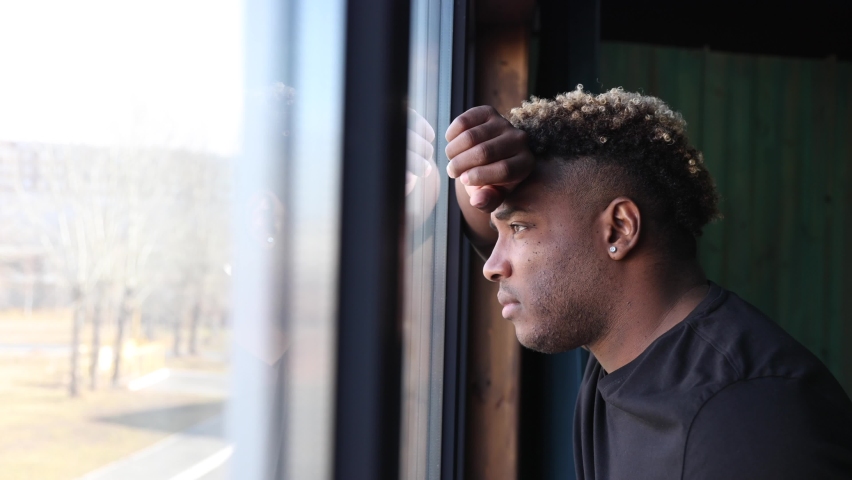 Portrait of a sad black man standing at the window. An African guy in a black T-shirt looks out the window, he is sad, he is depressed, there was a crisis in his life | Shutterstock HD Video #1088437561