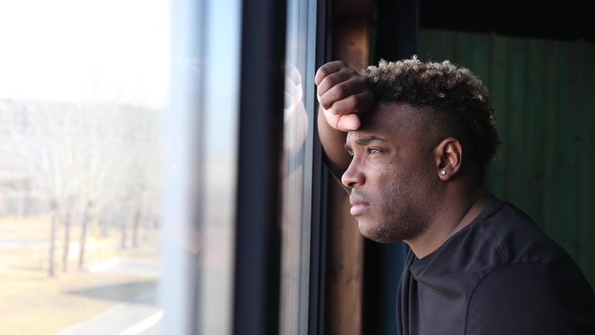 Portrait of a sad black man standing at the window. An African guy in a black T-shirt looks out the window, he is sad, he is depressed, there was a crisis in his life