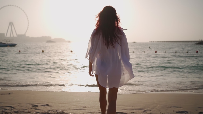 Woman in a white shirt walks along the beach of Persian Gulf in Dubai towards the sea against the backdrop of a beautiful sunset. Journey through the United Arab Emirates. Go Everywhere. Slow motion