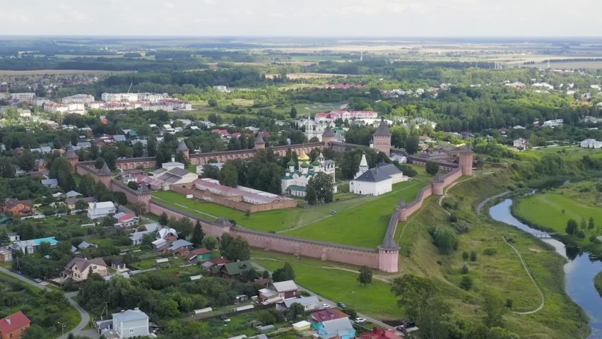 Suzdal, Russia. The Saviour Monastery of St. Euthymius, founded in 1352, is seen from above as a massive fortified monastic complex enclosed by tall red-brick walls with multiple towers. The monastery sits on a grassy rise beside a winding river, with the seven-domed Cathedral of the Transfiguration and other white-walled churches visible within the enclosure. The surrounding landscape is lush and green, with scattered houses, dense tree cover, and open fields stretching to the horizon under a partly cloudy summer sky. The camera executes a smooth dolly zoom that compresses the depth between the fortress walls and the distant town, emphasizing the monumentality of the 17th-century defensive architecture. The ensemble is part of the UNESCO World Heritage Site "White Monuments of Vladimir and Suzdal," and the scene captures the serene, historic atmosphere of one of Russia