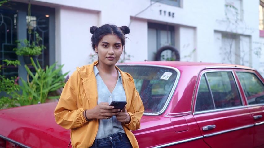 A young modern stylish retro smiling Indian Asian woman is standing outdoors leaning on a red vintage car and using a mobile phone in a city or urban setting looking in the camera. Fashionable trendy  Royalty-Free Stock Footage #1088438847