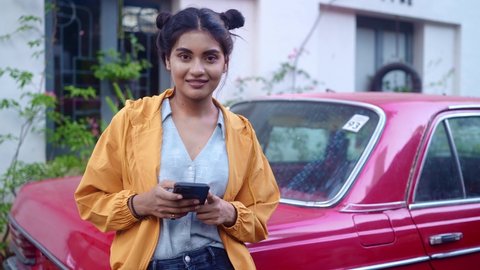 A young modern stylish retro smiling Indian Asian woman is standing outdoors leaning on a red vintage car and using a mobile phone in a city or urban setting looking in the camera. Fashionable trendy 