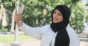 Happy muslim woman communicates by video call on street in summer. Woman smiles, waves her hand to telephone camera. Online video connection on city. Arab woman talking, communicating using phone