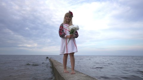 Wide shot happy little Ukrainian girl in traditional embroidered clothing standing on river pier with bouquet of flowers looking at camera smiling. Portrait of charming child posing in slow motion