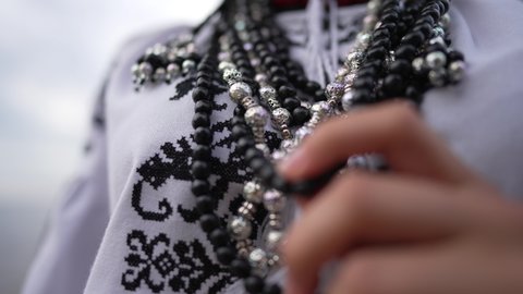 Close-up female hand touching traditional Ukrainian necklace on embroidered national shirt. Unrecognizable slim young woman in folk black and white clothing standing outdoors. Slow motion