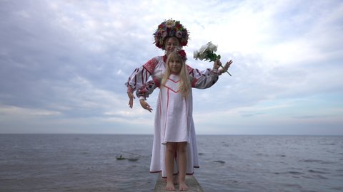 Wide shot portrait of confident happy Ukrainian daughter and mother in traditional embroidered dresses moving hands in slow motion standing on river pier. Smiling beautiful girl and woman posing