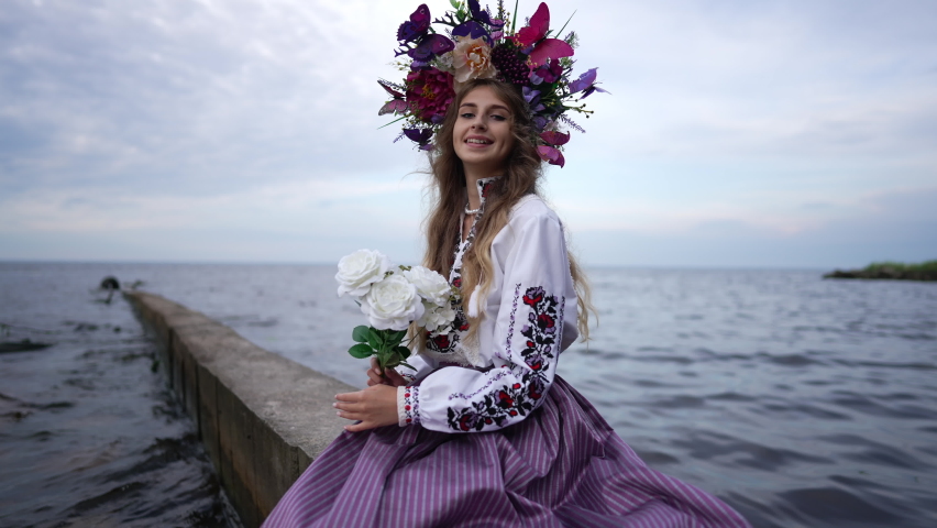 Smiling beautiful Ukrainian young woman with bouquet of flowers stretching hand looking at camera. Portrait of positive confident charming lady posing on river pier outdoors in slow motion