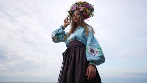 Young Ukrainian woman in embroidered shirt skirt and head wreath standing at background of cloudy sky. Portrait of slim beautiful lady in traditional clothing outdoors. Slow motion