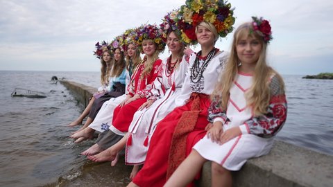 Wide shot positive Ukrainian women and girl sitting on river pier looking at camera smiling. Happy confident beautiful ladies posing outdoors in slow motion with cloudy summer spring sky at background