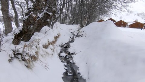 A small, partly frozen stream flowing down a snowy winter landscape with trees and some chalets in the background. Weather is snowy. Wintersport area.