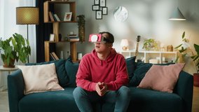 Asian man playing video games. Happy young Korean guy wearing virtual reality glasses, sitting on sofa in living room, male person enjoying VR 3D game. 