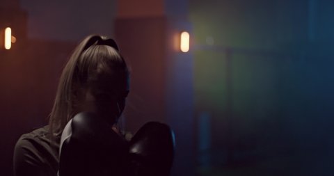 Portrait, a girl professionally practices punches in a dark hall of a fight club, boxing gloves on her hands. Silhouette, a girl boxer strikes in fighting gloves, a serious look of the face. Dark room