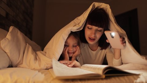 Family bedtime. Fairytale time. Pretty young mom and cute child daughter reading a book with a phone flashlight under the blanket in bed