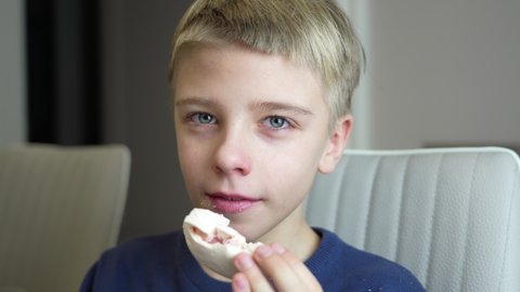 Nice happy boy eats meringue at home. Cute kid greedily eating big meringue at light kitchen