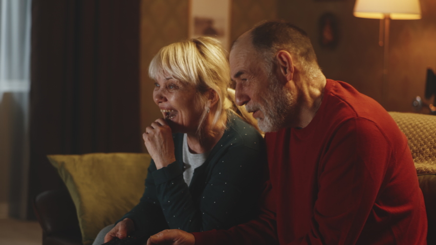 Happy senior husband and wife laughing at funny joke while sitting on couch and watching comedy on TV at night at home Royalty-Free Stock Footage #1088456265