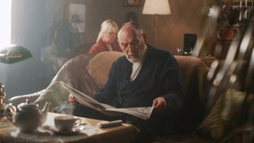 Pan left view of aged man in bathrobe and glasses sitting on couch and reading daily newspaper in cozy living room, near woman in morning at home - Powered by Shutterstock - Get 15% off with code: PIKWIZARD15