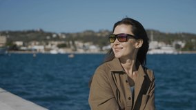 A happy girl, a brunette, in sunglasses, of Asian appearance, sits on a pier on the seashore, smiles. The wind blows your hair, the sun shines. Medium plan.