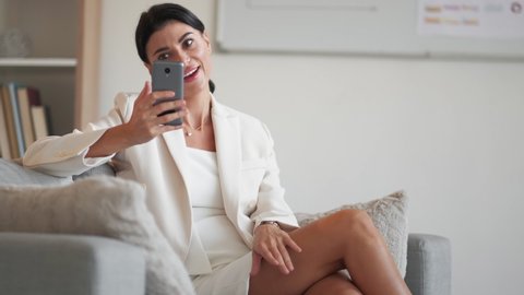 Virtual meeting. Coquettish woman. Luxury lifestyle. Happy business lady in stylish suit dress talking affectedly on smartphone video call sitting couch in light room interior.