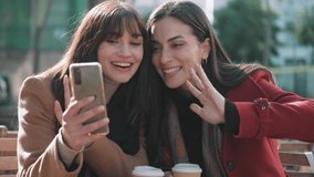 Video of two attractive friends enjoying coffee together while doing a videocall with smartphone sitting on the terrace of a bar.