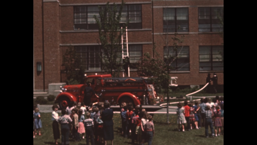 1950s: firemen in fire hats hold hose, water shoots from hose, kids jump near school and fire truck as fireman climbs down ladder, water reverses into hose held by firemen, "fire drill" on slate