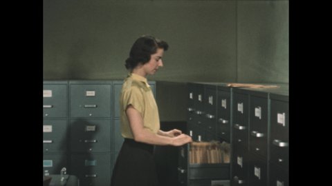 1950s: File cabinet drawer. Hands tab to the January tab and pull a folder. Folder is replaced with an Out tab and slip. Secretary closes the drawer. She lectures.