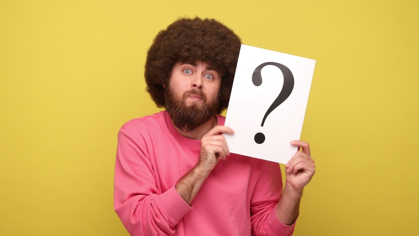 Bearded hipster puzzled man with Afro hairstyle looking at camera, holding paper with question mark, thinks about tasks, wearing pink sweatshirt. Indoor studio shot isolated on yellow background.