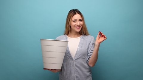 Portrait of attractive woman with wavy hair holding white trash can and throwing out her eyeglasses after laser eyes treatment, wearing striped shirt. Indoor studio shot isolated on blue background.