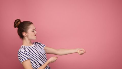Persistent woman pulling invisible rope with great effort, being purposeful, using all strength and energy to achieve goal, wearing striped T-shirt. Indoor studio shot isolated on pink background.