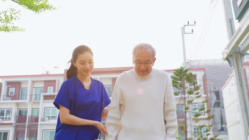 Asian young caregiver nurse support senior older male walking outdoors. Specialist girl doctor help and take care of elderly mature man patient doing physical therapy in public park at nursing home. Royalty-Free Stock Footage #1088471717