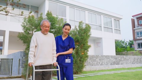 Asian young caregiver nurse support senior older male walking outdoors. Specialist girl doctor help and take care of elderly mature man patient doing physical therapy in public park at nursing home.
