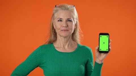 Smiling mature woman posing holding phone with tracking points on blank empty green screen, isolated on orange background. Giving mahalo shaka hand sign signal saying hello. 
