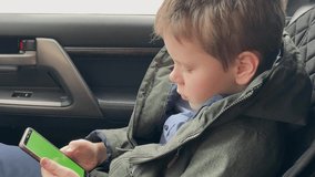 4K Boy dressed in a winter sweatshirt sitting in the car and playing mobile game on smartphone holding smartphone with green screen