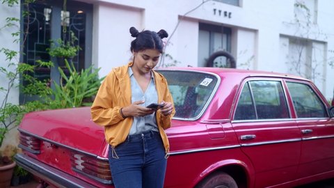 A young modern stylish retro smiling Indian Asian woman is standing outdoors leaning on a red vintage car and using a mobile phone in a city or urban setting. Concept of fashion, trend, and technology Vídeo Stock