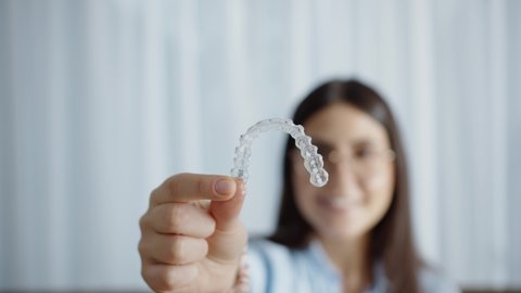 Happy Attractive Woman Smiles And Sitting In Cozy House And Holding Invisalign Braces, Transparent Plastic Retainers Or Tooth Whitening System. Concept Of Dental Healthcare And Orthodontic