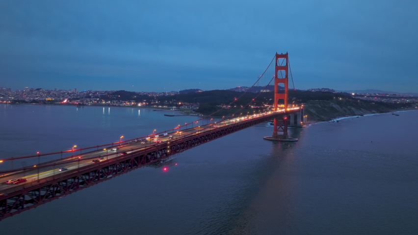 Establishing aerial view shot of San Francisco City Skyline in distance and illuminated Red Golden Gate Bridge in foreground. San Francisco downtown on background under deep blue gray clouds at sunset Royalty-Free Stock Footage #1088480333