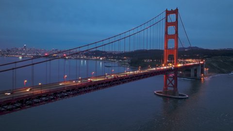 Establishing aerial view shot of San Francisco City Skyline in distance and illuminated Red Golden Gate Bridge in foreground. San Francisco downtown on background under deep blue gray clouds at sunset