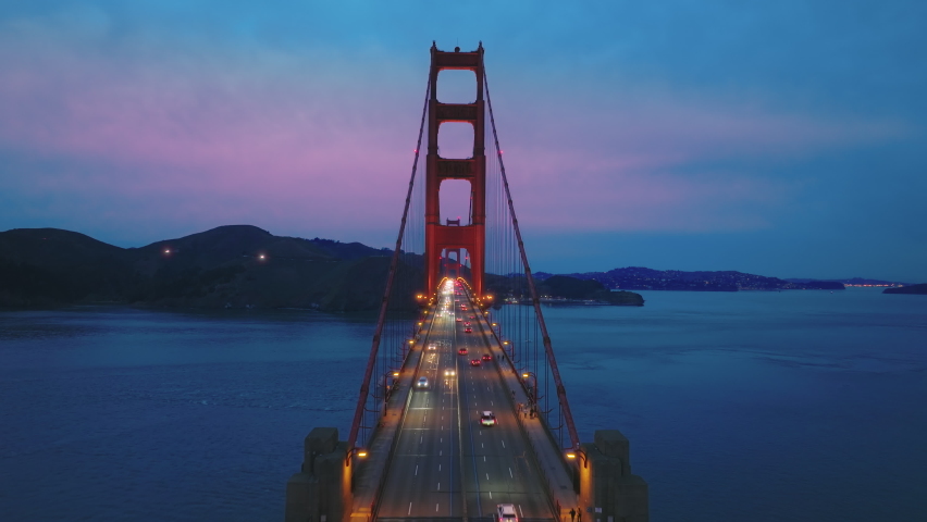 Aerial video of the Golden Gate Bridge at night. Inspirational drone flight through red tower above busy traffic road. Cinematic pink cloudy sunset over San Francisco bay on background, California USA