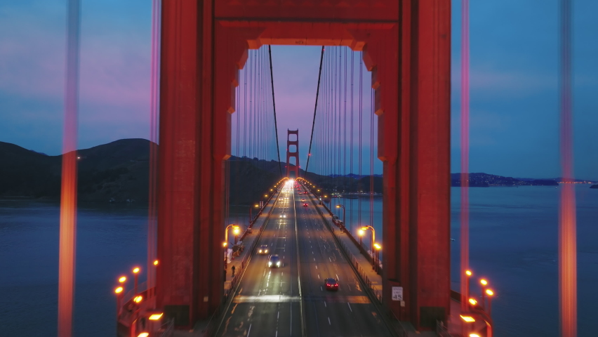 Aerial video of the Golden Gate Bridge at night. Inspirational drone flight through red tower above busy traffic road. Cinematic pink cloudy sunset over San Francisco bay on background, California USA