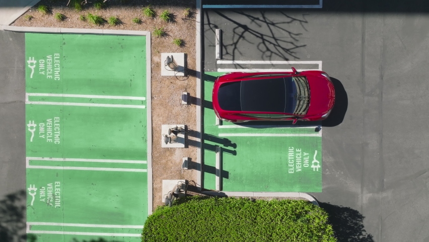 Aerial of electric car vehicles recharging. EV eco friendly electricity powers cars. Drone descending above man plugging in charging cord in red SUV sedan electric vehicle at summer, RED camera Royalty-Free Stock Footage #1088482017